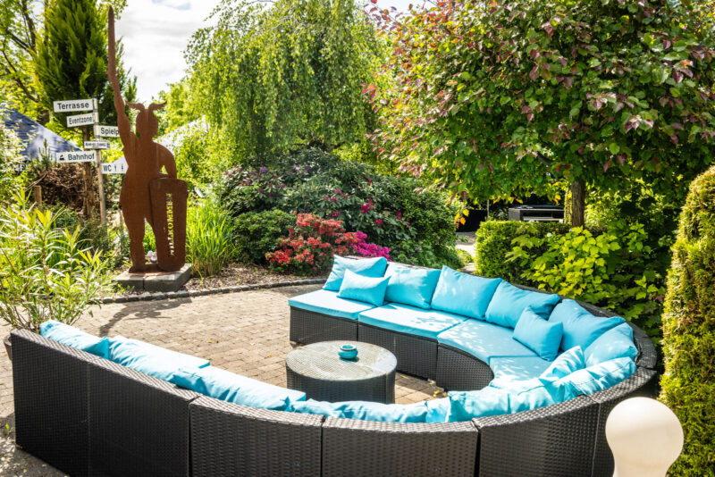 Halbrundes Rattan-Loungesofa mit türkisfarbenen Kissen und Willkommens-Skulptur im Außenbereich des Landhaus Begatal
