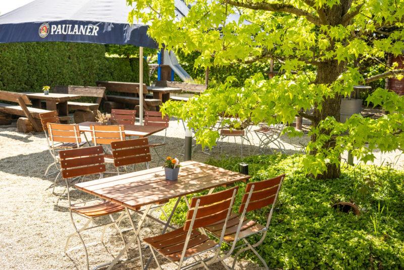 Sonniger Biergarten des Landhaus Begatal mit Holztischen, Bistrostühlen und Paulaner-Sonnenschirmen unter einem Ahornbaum