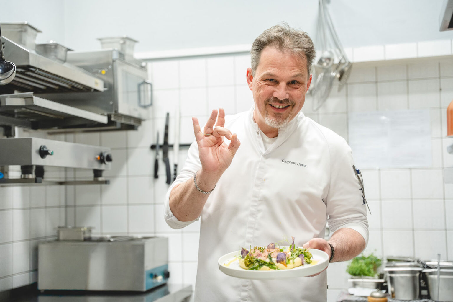 Küchenchef Stephan Büker präsentiert lachend einen angerichteten Teller mit OK-Handzeichen in der Küche des Landhaus Begatal