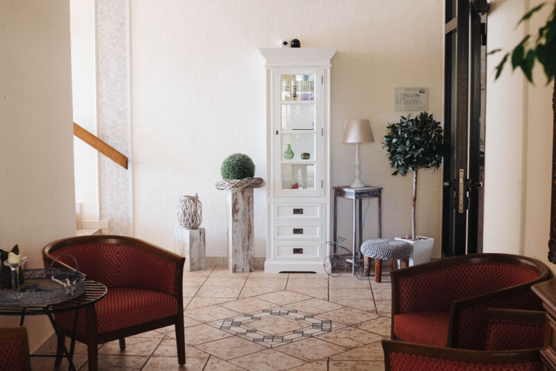 Einladende Hotellobby des Landhaus Begatal mit roten Loungesesseln, weißem Vitrinenschrank und dekorativen Zimmerpflanzen