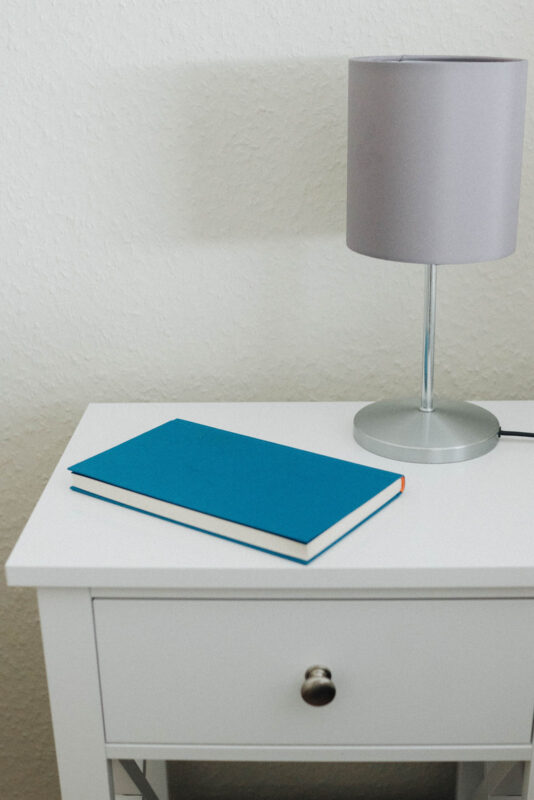 Weißer Nachttisch mit Nachttischlampe und blauem Buch im Hotelzimmer des Landhaus Begatal