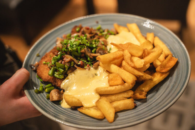 Schnitzel mit Pommes, cremiger Sauce, Pilzen, Speck und grünen Bohnen auf einem blauen Teller.